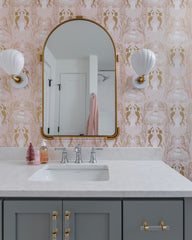 This elegant bathroom showcases the white Coquille sconce, a shell-shaped wall fixture, paired with whimsical gold mermaid wallpaper, a marble vanity, and a gold-trimmed mirror. The combination of soft tones and gold accents creates a refined coastal-inspired look.