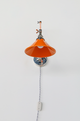 An orange plug-in wall lamp with a glossy cone shade and polished nickel hardware, shown from the front so you can see the bulb, the swivel joints, and the gray twisted cloth cord with its inline switch. The cord plugs into any outlet, so you can put a sconce where there is no junction box. Hang one on each side of a bed or beside a reading chair. It works well in rentals, apartments, and kids rooms, and fits retro, industrial, and colorful modern decor.