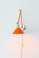 A plug-in swing arm wall sconce with an orange cone shade, orange arms, and nickel joints, shown lit with the arm folded close to the wall. A gray twisted cloth cord runs down from the backplate to an inline on/off switch, so there is no hardwiring and no electrician. Mount it by a bed as a reading light or over a desk. Bright orange and polished nickel look sharp on white or pale gray walls, so it suits renter friendly bedrooms, dorms, and colorful mid century rooms.