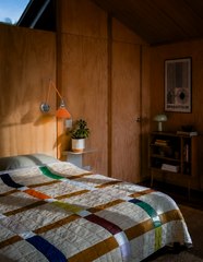 Warm plywood paneling and an angled cabin ceiling set the tone in this retro bedroom, where a Bauhaus exhibition poster and a ceramic mushroom lamp do the talking on style. The real hero is the Cora Sconce in Orange and Chrome, its double-jointed swing arm and exposed wingnut hardware reading like vintage drafting equipment scaled up for the bedroom. A rainbow-striped patchwork quilt keeps the whole room from feeling too serious. Mid-century cabin bedroom inspiration for anyone who wants their wood paneling