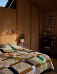 Warm plywood paneling and an angled cabin ceiling set the tone in this retro bedroom, where a Bauhaus exhibition poster and a ceramic mushroom lamp do the talking on style. The real hero is the Cora Sconce in Orange and Chrome, its double-jointed swing arm and exposed wingnut hardware reading like vintage drafting equipment scaled up for the bedroom. A rainbow-striped patchwork quilt keeps the whole room from feeling too serious. Mid-century cabin bedroom inspiration for anyone who wants their wood paneling