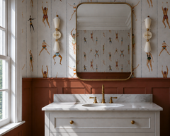 Playful traditional bathroom with cream and brass Janus sconces flanking a large brass-framed mirror over a marble vanity. Vintage swimmer wallpaper, terra cotta paneled wainscoting, brass faucet, cream cabinetry, and classic molding give the room a polished but quirky grandmillennial feel. The geometric two-light sconces add sculptural personality and work beautifully for a kids bathroom, powder room, or maximalist bath with vintage sports wallpaper and warm brass vanity lighting.