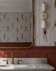 This corner view catches the terracotta and cream powder room in real morning light, sun raking across the wallpaper's rows of vintage-style bathers and warming up the wainscoting below. The Cream Janus sconce sits just off the mirror's edge, its rounded cone shades and brass stem picking up the same warmth as the light. A small soap dish and sponge on the marble counter add the kind of lived-in detail that makes a powder room feel like part of a real house.