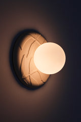 Lit Lierre ceramic wall sconce in unglazed cream porcelain, botanical leaf relief backplate with matte white globe diffuser, warm ambient glow for bedroom or entryway.