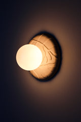 Lit Lierre ceramic wall sconce in unglazed cream porcelain, botanical leaf relief backplate with matte white globe diffuser, warm ambient glow for bedroom or entryway.