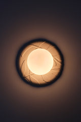 Front view of lit ceramic globe wall sconce, unglazed cream porcelain leaf relief plate surrounding matte white globe shade, minimalist flush mount style light.