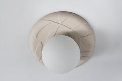 Lierre ceramic wall sconce or ceiling light in unglazed cream porcelain, sculptural leaf relief plate with matte white globe diffuser, modern botanical lighting on white wall.