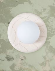 Close-up product styling image of the Cream Lierre ceramic wall sconce by Sazerac Stitches, featuring a soft cream glazed, leaf-veined ring shade surrounding a matte white globe diffuser. The fixture is photographed against muted sage green lichen-inspired botanical wallpaper for an earthy, organic modern look that feels calm and natural. Ideal visual for SEO keywords like ceramic wall sconce, cream ceramic lighting, botanical wallpaper, sage green decor, organic modern interior, and nature-inspired lightin