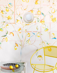 Bright, airy sunroom vignette featuring a Cream Lierre ceramic wall sconce by Sazerac Stitches mounted on whimsical lemon-and-vine wallpaper with a soft blush grid pattern. A bold yellow metal lounge chair adds a punch of color, paired with a glass-topped side table holding a modern ceramic tray and fresh lemons. The overall decor feels Mediterranean coastal meets playful modern maximalism, with cheerful citrus colors, hand-painted botanical wallpaper, and a light, happy summer-at-home vibe.