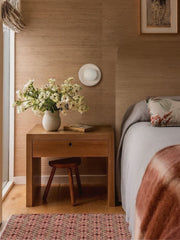 Cream matte porcelain Lierre ceramic wall sconce with leafy relief texture and frosted globe light, installed above a wood nightstand in a warm neutral bedroom with tan grasscloth wallpaper, vintage inspired bedding, and soft natural light, handcrafted small batch lighting designed and assembled in New Orleans with ceramic cast and glazed in North Carolina, organic modern coastal traditional bedroom wall lighting