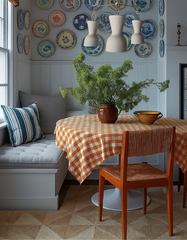 Inviting breakfast nook featuring the Cream & Brass Margot chandelier with sculptural ceramic shades, hanging above a round table covered in an orange gingham tablecloth. The corner includes gray tufted banquette seating, a mid-century wood chair, blue-and-white decorative wall plates, and a greenery centerpiece, blending artisanal handmade lighting with traditional cottage-inspired design.