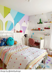 Wide view of a pastel teenage girls bedroom with a white metal bed, rainbow bedding, geometric mural wall, white desk with pink daisy drawer pulls, pink textured desk chair, and built-in shelving for toys and display pieces. Bright eclectic girls bedroom with playful color, flower drawer hardware, feminine bedroom decor, tween girl room ideas, pastel interior design, creative kids room styling, and custom desk details that add personality.