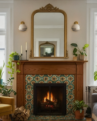 Pair of Delta ceramic globe sconces with brass caps and crisp black stripe flank an ornate gold mirror above a wood mantel, highlighting a glowing tiled fireplace with green patterned surround, stacked firewood, taper candles, and layered houseplants in an inviting, collected living room.