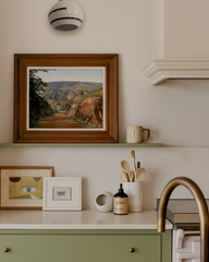 Delta ceramic globe sconce with bold black stripes mounted above a softly layered kitchen vignette featuring sage green cabinetry with brass knobs, cream walls, framed landscape and abstract art, slim display shelf, ceramic mug, minimalist utensil crock, sculptural match striker, brass gooseneck faucet, and stainless range, creating a warm modern-cottage feel.