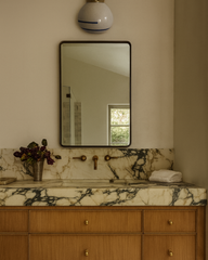 Single Delta ceramic globe sconce with black stripe and brass fitter centered above a tall mirror in a warm neutral bathroom, featuring European-inspired marble backsplash and thick veined marble countertop, minimalist brass wall-mount faucet, light wood vanity with brass knobs, folded white towel, and moody florals in a metal cup.