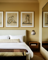 Single Delta ceramic globe sconce with brass cap and black accent stripe mounted above a wood nightstand in a warm camel-toned bedroom, styled with a low upholstered bed in pale cream linen, layered white bedding and brown blanket, olive velvet bench, and trio of framed nude line drawings, creating a soft, modern, gallery-inspired retreat.