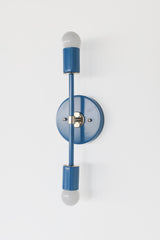 Close angled studio photo of the Denim Blue Medium Thalia Sconce with chrome hardware and two white globe bulbs. The glossy blue finish adds color while the chrome details keep the fixture feeling crisp and clean. This vertical wall sconce is ideal for bathroom vanity lighting, small powder rooms, playful kids bathrooms, modern coastal spaces, and colorful custom interiors.