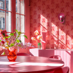 A pink shell sconce in a sun-drenched, all-pink Parisian room — pure pink-on-pink drama. The Coquille from Sazerac Stitches is a glossy scallop-shell wall light with a warm brass fitting, in a doll-pink hue that sits right into a palm-tree wallpaper in red and blush. Tall shuttered windows, a rounded pink table, sculptural pink vases, and a loose spring bouquet complete the color-drenched scene. Romantic, feminine, and unapologetically monochromatic. Works for pink room ideas, color-drenched interiors, palm