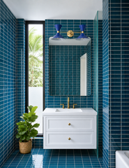 A two-light vanity light with cobalt blue double cone shades on a brass bar, mounted above a rounded mirror. The bathroom is wrapped floor to ceiling in glossy blue vertical stack tile with white grout, with a white floating shaker vanity, brass faucet and knobs, a tall narrow window onto palm trees, and a fiddle leaf fig in a woven basket. Blue on blue with brass reads tropical and modern, a good fit for a coastal bathroom, mid century modern bath, or blue tile powder room.