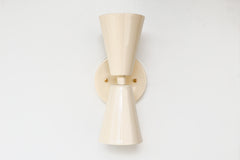 Close-up of the Cream Dryades Sconce, a double-cone wall light fixture handcrafted in powder-coated steel with a soft cream finish and subtle sheen. This mid-century modern-inspired wall sconce features two symmetrical cones joined at a central brass-accented baseplate, creating a sculptural and timeless silhouette. Ideal for bathrooms, hallways, and bedrooms, this handcrafted light fixture adds warmth and minimal elegance to any space. Shown against a clean white background to highlight the glossy neutral 