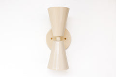 Close-up of the Cream Dryades Sconce, a double-cone wall light fixture handcrafted in powder-coated steel with a soft cream finish and subtle sheen. This mid-century modern-inspired wall sconce features two symmetrical cones joined at a central brass-accented baseplate, creating a sculptural and timeless silhouette. Ideal for bathrooms, hallways, and bedrooms, this handcrafted light fixture adds warmth and minimal elegance to any space. Shown against a clean white background to highlight the glossy neutral 