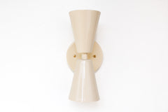 Close-up of the Cream Dryades Sconce, a double-cone wall light fixture handcrafted in powder-coated steel with a soft cream finish and subtle sheen. This mid-century modern-inspired wall sconce features two symmetrical cones joined at a central brass-accented baseplate, creating a sculptural and timeless silhouette. Ideal for bathrooms, hallways, and bedrooms, this handcrafted light fixture adds warmth and minimal elegance to any space. Shown against a clean white background to highlight the glossy neutral 