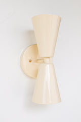Close-up of the Cream Dryades Sconce, a double-cone wall light fixture handcrafted in powder-coated steel with a soft cream finish and subtle sheen. This mid-century modern-inspired wall sconce features two symmetrical cones joined at a central brass-accented baseplate, creating a sculptural and timeless silhouette. Ideal for bathrooms, hallways, and bedrooms, this handcrafted light fixture adds warmth and minimal elegance to any space. Shown against a clean white background to highlight the glossy neutral 