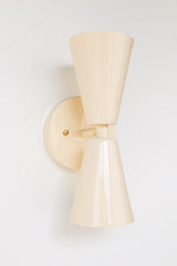 Close-up of the Cream Dryades Sconce, a double-cone wall light fixture handcrafted in powder-coated steel with a soft cream finish and subtle sheen. This mid-century modern-inspired wall sconce features two symmetrical cones joined at a central brass-accented baseplate, creating a sculptural and timeless silhouette. Ideal for bathrooms, hallways, and bedrooms, this handcrafted light fixture adds warmth and minimal elegance to any space. Shown against a clean white background to highlight the glossy neutral 