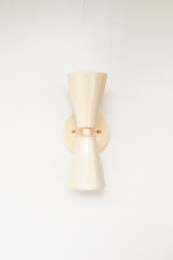 Close-up of the Cream Dryades Sconce, a double-cone wall light fixture handcrafted in powder-coated steel with a soft cream finish and subtle sheen. This mid-century modern-inspired wall sconce features two symmetrical cones joined at a central brass-accented baseplate, creating a sculptural and timeless silhouette. Ideal for bathrooms, hallways, and bedrooms, this handcrafted light fixture adds warmth and minimal elegance to any space. Shown against a clean white background to highlight the glossy neutral 