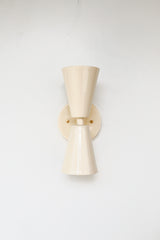 Close-up of the Cream Dryades Sconce, a double-cone wall light fixture handcrafted in powder-coated steel with a soft cream finish and subtle sheen. This mid-century modern-inspired wall sconce features two symmetrical cones joined at a central brass-accented baseplate, creating a sculptural and timeless silhouette. Ideal for bathrooms, hallways, and bedrooms, this handcrafted light fixture adds warmth and minimal elegance to any space. Shown against a clean white background to highlight the glossy neutral 