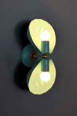 Double Coquille sconce in poolside teal with brass hardware, featuring two fluted, scalloped shell-shaped shades mounted top and bottom on a round teal backplate with an ornate brass floral rosette at the center. Both exposed bulbs are lit, casting a warm glow through the shell shades against a dark charcoal wall, showing the coastal, grandmillennial-style silhouette of the fixture.