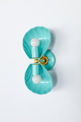 Double Coquille sconce in poolside turquoise with brass hardware, straight-on front view showing two fluted, scalloped shell-shaped shades mounted top and bottom on a round poolside backplate with an ornate brass floral rosette at the center, unlit with exposed white bulbs.