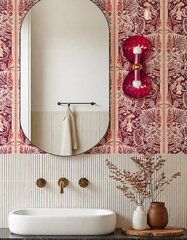 A colorful, design-forward bathroom featuring a tall rounded mirror, creamy fluted wall tile, a sculptural white sink, warm brass wall-mounted faucet, and peach-and-burgundy wallpaper with a whimsical mermaid and seaside toile feel. The star of the space is a glossy berry pink double shell sconce with brass detailing, adding a playful coastal glam vibe that feels equal parts boutique hotel, girls bathroom, and elevated powder room. This look is perfect for anyone searching colorful bathroom ideas, shell sco