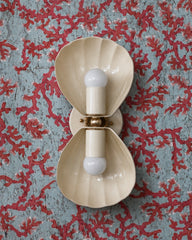 Ivory two-light wall sconce with fluted shell shades and a brass rosette center, mounted vertically. The cream color stays quiet, so it can handle a loud wall, here a pale blue wallpaper covered in red branching coral. Suits a coastal grandmillennial bathroom, a Palm Beach style powder room, or a vanity light beside a beach house mirror. Cream, coral red, and powder blue with warm brass is a classic seaside palette. Handmade in New Orleans in 30 colors.