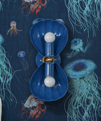 Denim blue two-light wall sconce with fluted shell shades, a brass rosette, and a brass ball center, mounted vertically. It sits on dark navy wallpaper with glowing aqua, violet, and coral jellyfish, so the shells almost melt into the deep-sea print. A strong pick for a moody blue bathroom, a dark powder room, or an ocean-themed kids' bath adults will like too. Blue on blue with brass keeps it rich instead of beachy. Handmade in New Orleans in 30 colors.