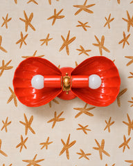 Red-orange two-light wall sconce with fluted shell shades, mounted horizontally so it works as a vanity light above a mirror. Brass rosette and ball at the center. Shown on cream wallpaper scattered with tan starfish, a warm, sunny take on coastal decor. Fits a playful beach house bathroom, a colorful powder room, or a retro seaside kitchen. The red pops hard against a neutral wall, so let it be the one bright thing in a calm room. Handmade in New Orleans in 30 colors.