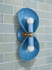 Side view of a blue two-light wall sconce with deep fluted shell shades that curve around each bulb, set off the wall on a round backplate with a brass rosette and ball. Mounted on pale aqua subway tile with white grout, it works as a bathroom vanity light or a sconce over a kitchen backsplash. Blue on aqua keeps the palette soft and monochromatic, with brass for warmth. A coastal spa bathroom idea with a little art deco curve. Handmade in New Orleans in 30 colors.