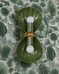 Olive green two-light wall sconce with fluted shell shades, a brass rosette, and a brass ball center, mounted vertically. Shown on pale gray wallpaper printed with green fan coral and seaweed, so the whole wall reads green and earthy. A good fit for a moody green bathroom, a vintage botanical powder room, or a cottage-style vanity light. Olive takes the shell shape somewhere less beachy and more old naturalist's study. Handmade in New Orleans in 30 colors.