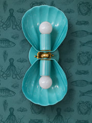 Turquoise two-light wall sconce with fluted shell shades, a brass rosette, and a brass ball at the center, mounted vertically. Shown on teal wallpaper printed with vintage-style octopus, crabs, fish, and jellyfish in a darker teal, so the sconce reads as a brighter version of the wall. Good for a nautical powder room, coastal bathroom vanity, or moody beach house hallway. Tonal teal with brass is an easy way to go bold without clashing. Handmade in New Orleans in 30 colors.