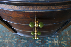 A close-up look at a brass and olive green double-ball knob installed on the bowed drawer front of a dark, aged wood nightstand. The glossy green spheres stand out against the wood's deep patina, showing how one colorful hardware swap can update an inherited or secondhand piece without a full refinish. A worn, vintage-toned rug underneath keeps the whole scene feeling collected rather than new.