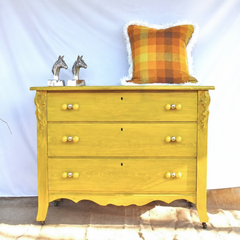 A vintage dresser painted a confident mustard yellow, with carved scroll details, a scalloped skirt, and small casters, styled with silver horse head bookends and a warm plaid pillow. Double-ball yellow knobs finish off each of the three drawers, tying the hardware back into the paint color instead of defaulting to something plain. A good before-and-after reference for anyone repainting an inherited or secondhand dresser.