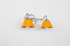 Angled view of the Double Kelly Sconce in matte mustard yellow with polished chrome hardware, a compact double vanity light with powder coated steel cone shades and exposed white globe bulbs. Great for small bathrooms, powder rooms, or paired over each mirror in larger bathrooms, this colorful wall fixture adds personality, retro charm, and modern eclectic style.