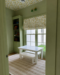 Color-drenched pastel green playroom with a lemon-print ceiling and matching Roman shade, built-in storage, and a kid-sized white table and bench on a natural jute rug. A spring green and brass double wall sconce adds playful, modern cottage lighting. Great for playroom design, nursery lighting, lemon wallpaper ceiling, color drenched interiors, and brass accent lighting.