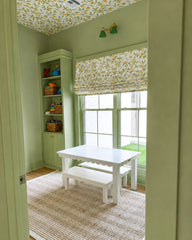 Color-drenched pastel green playroom with a lemon-print ceiling and matching Roman shade, built-in storage, and a kid-sized white table and bench on a natural jute rug. A spring green and brass double wall sconce adds playful, modern cottage lighting. Great for playroom design, nursery lighting, lemon wallpaper ceiling, color drenched interiors, and brass accent lighting.