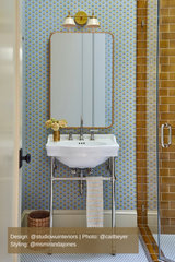 Charming bathroom featuring a mid-century modern brass and cream double-light sconce, blue and yellow patterned wallpaper, elegant brass-framed mirror, pedestal sink with chrome legs, woven wastebasket, and a tiled shower with rich topaz-colored subway tiles, creating a vibrant yet timeless interior.
