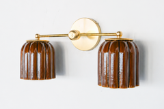 Handmade Double Marcella Sconce in chicory brown glazed ceramic — two fluted, scalloped shades on a brass arm and round backplate. Celadon-style glazing at its best: the glaze pools espresso-dark in the flutes and breaks to honeyed amber on the ribs, so it reads as a rich tortoiseshell neutral that turns jewel-toned in the light, not flat brown. Deco-inspired ribbing keeps it feminine and architectural. Layers beautifully with wood, cream, and leather. Shop handmade ceramic sconces, glazed brown wall lighti