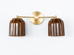 Handmade Double Marcella Sconce in chicory brown glazed ceramic — two fluted, scalloped shades on a brass arm and round backplate. Celadon-style glazing at its best: the glaze pools espresso-dark in the flutes and breaks to honeyed amber on the ribs, so it reads as a rich tortoiseshell neutral that turns jewel-toned in the light, not flat brown. Deco-inspired ribbing keeps it feminine and architectural. Layers beautifully with wood, cream, and leather. Shop handmade ceramic sconces, glazed brown wall lighti