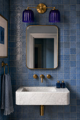 Bathroom vanity styled with the Double Marcella Sconce featuring two ribbed ceramic shades in a glossy cobalt blue glaze paired with a warm brass backplate and arms. Installed above a marble sink and gold-framed mirror against vibrant blue square tiles, this handmade wall light combines bold color, sculptural detail, and timeless craftsmanship. Designed by Sazerac Stitches in New Orleans, the double sconce is ideal for bathroom vanities, powder rooms, or modern eclectic interiors seeking colorful ceramic li