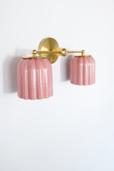 Angled view of a brass double wall sconce with two pastel pink glazed ceramic ribbed shades, a handmade ceramic vanity light with Art Deco lines and grandmillennial charm, ideal for powder rooms, bathrooms, and designer interiors mixing traditional decor with modern color