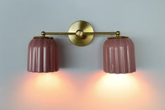 Double Marcella wall sconce turned on, featuring two glossy pastel pink ribbed ceramic shades with warm brass fixture, casting a soft ambient glow on the wall, a perfect ceramic vanity light for bathrooms and powder rooms, ideal for color lovers, chinoiserie rooms, and playful grandmillennial interiors